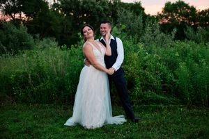 A romantic wedding couple poses together at sunset in a lush green field with trees in the background.