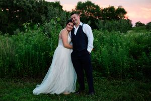 Newlyweds share an intimate moment at dusk in a natural outdoor setting with greenery and pink sky.