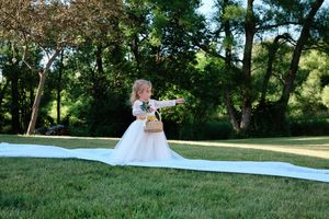 A couple in wedding attire poses for photos on a grassy hillside surrounded by trees on a sunny day.