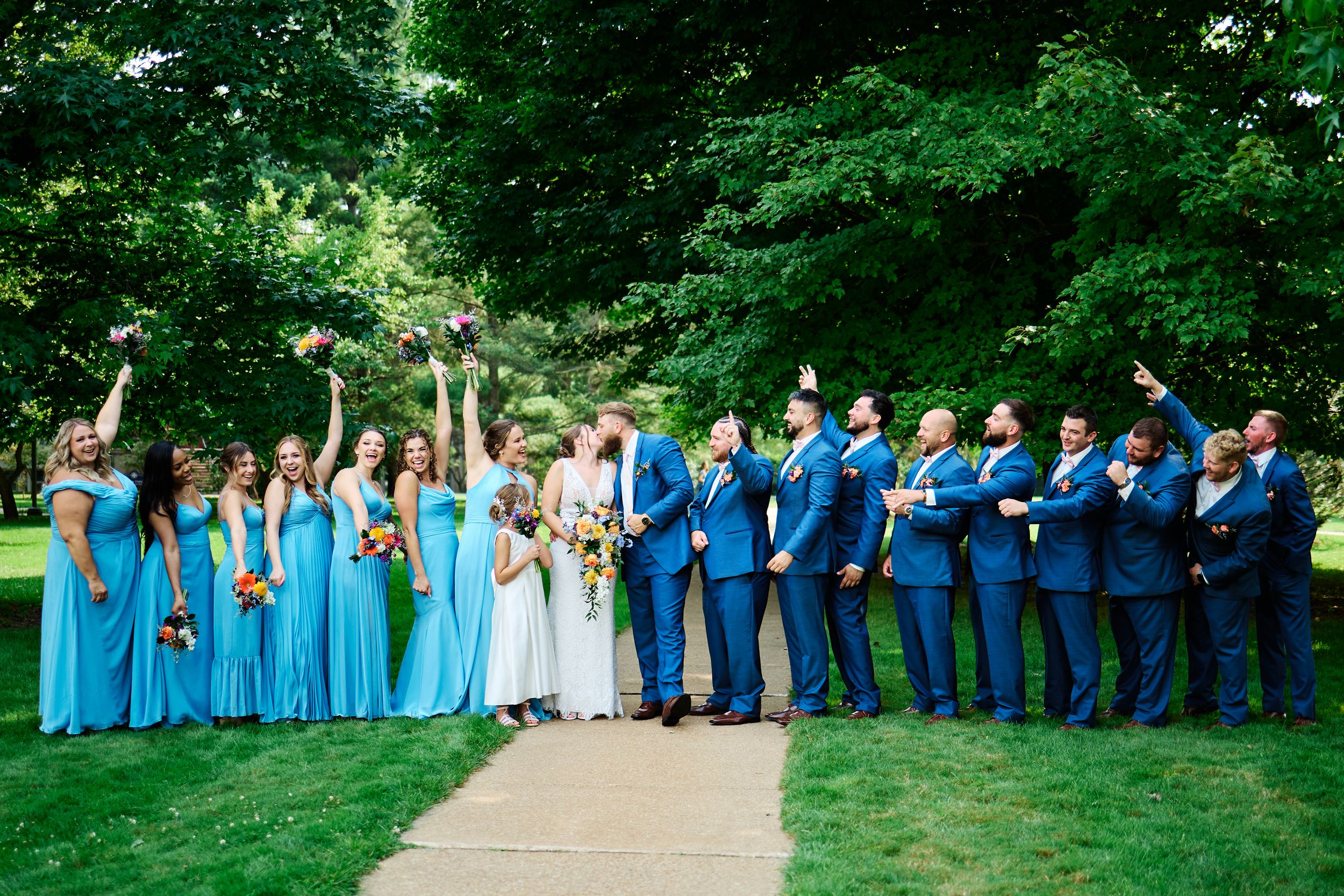 Wedding party in blue and navy attire poses together on a lush green lawn during a summer celebration.