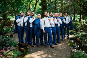 Wedding party in navy suits and bow ties pose together on garden path surrounded by lush greenery.