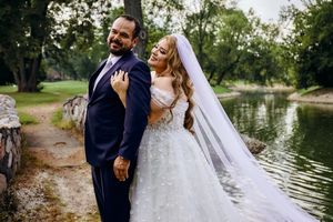 Romantic wedding couple embraces near a tranquil lake with a flowing white veil and trees in the background.