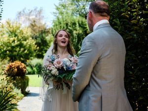 A couple in wedding attire embraces outdoors in front of lush greenery during their wedding ceremony.
