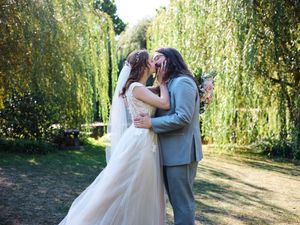 A couple in wedding attire embraces under cascading willow trees in a romantic garden setting.