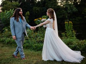 A couple in wedding attire walks through a garden setting during sunset while holding hands.