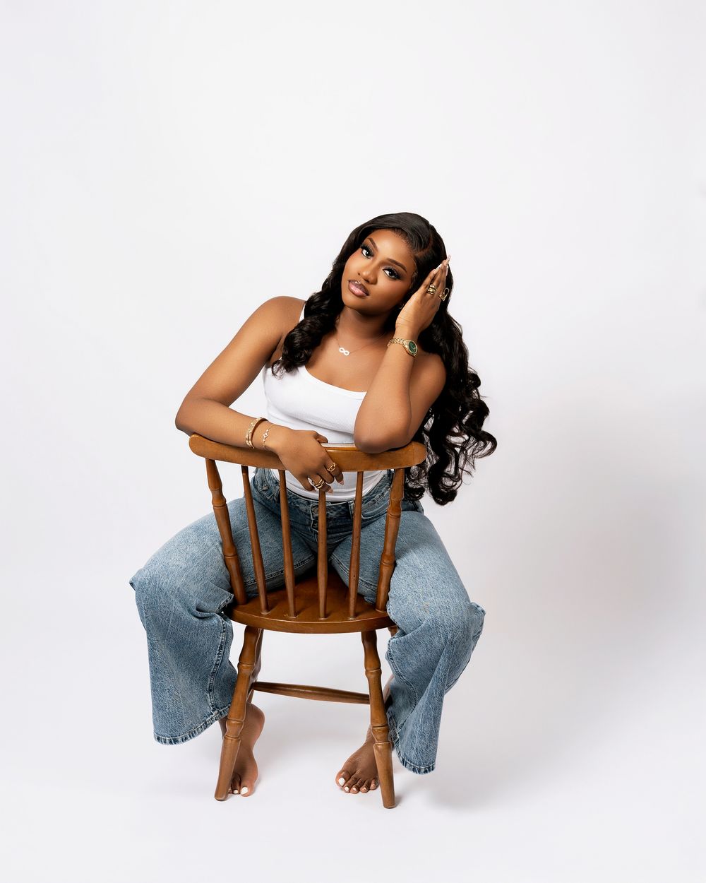 Young woman in white top and jeans poses gracefully on wooden chair against white background.