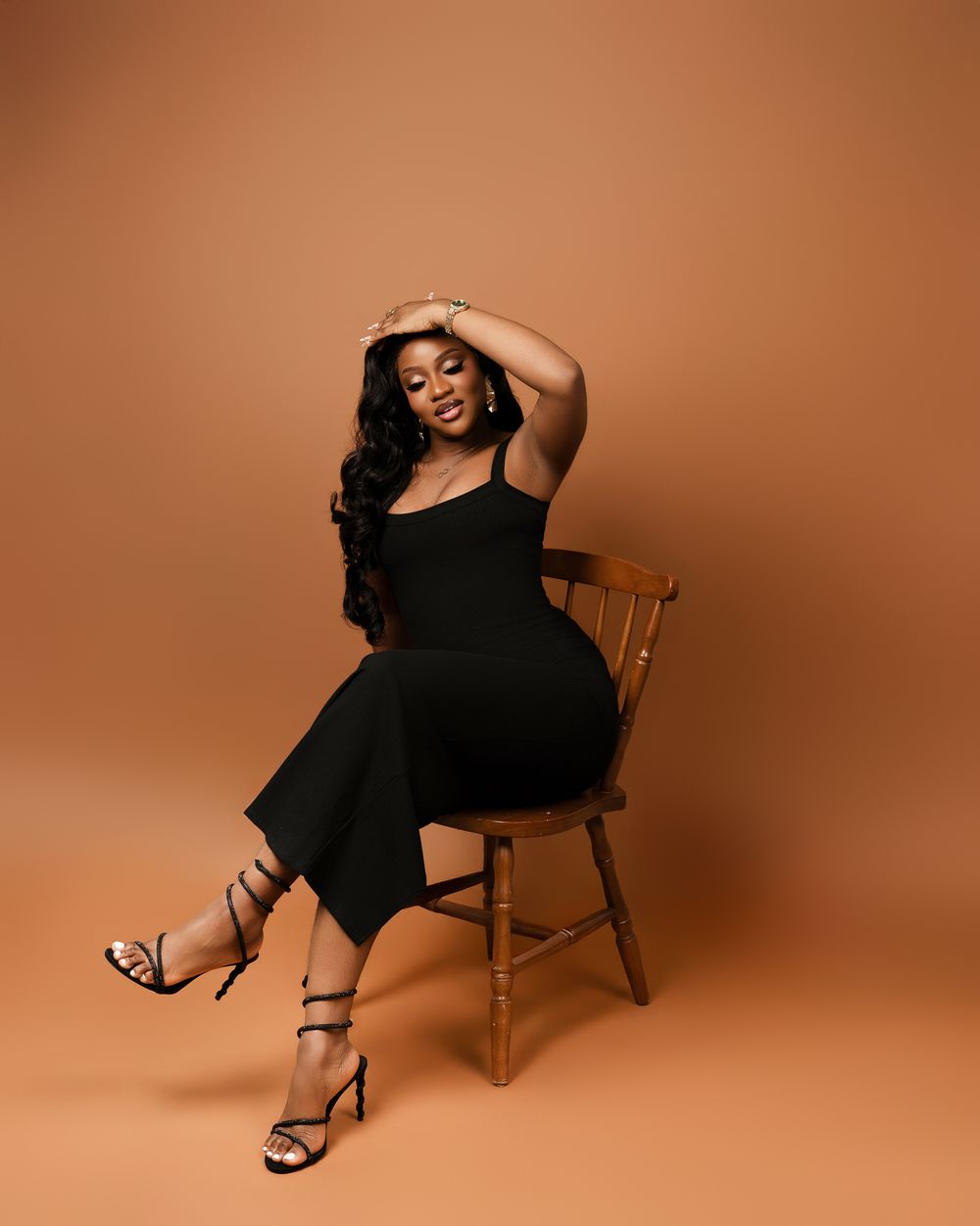 A woman in a black jumpsuit and strappy heels poses gracefully on a wooden chair against a warm tan background.