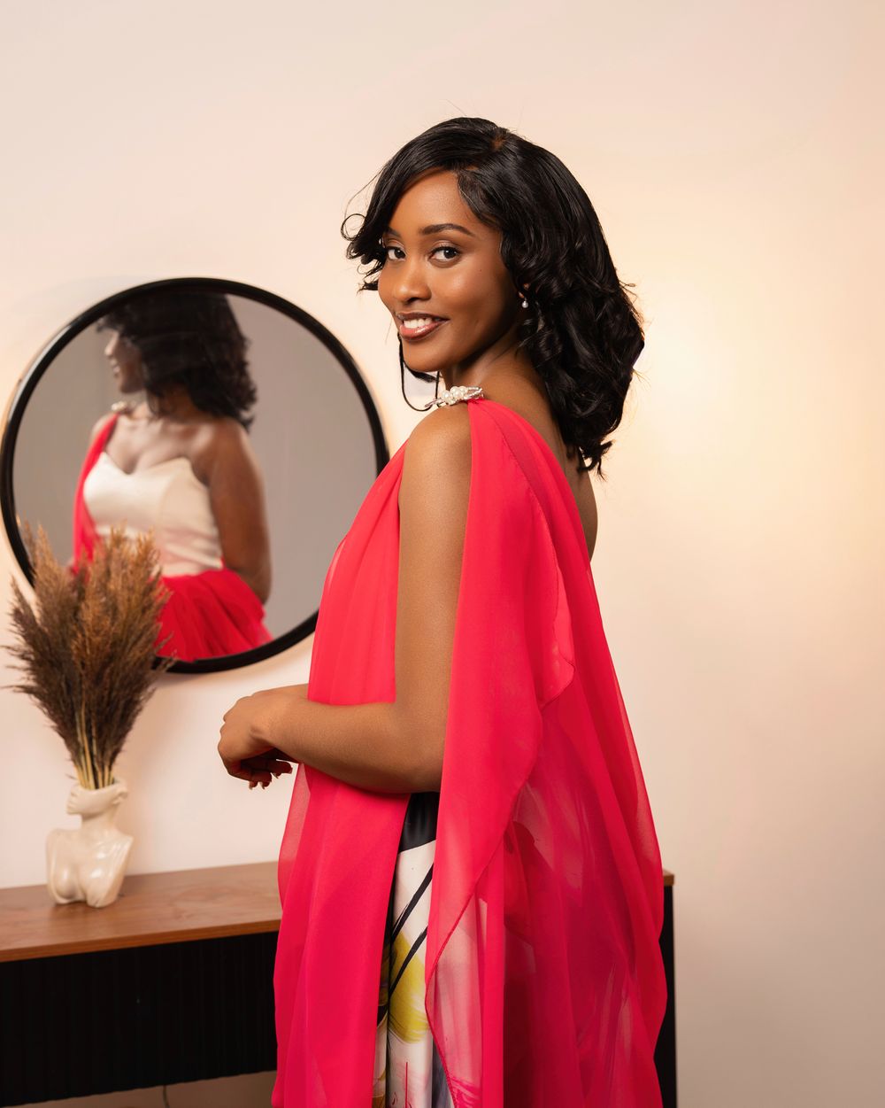 Woman in red chiffon cape dress smiling over shoulder, mirror reflection visible, elegant indoor setting.