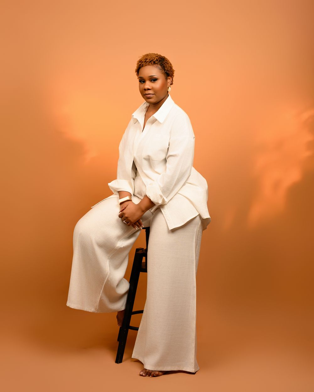 Studio portrait of someone in an elegant all-white ensemble seated on a stool against a warm orange backdrop.