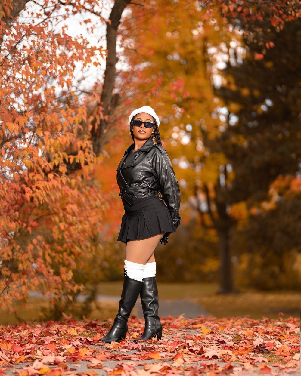 A person in black clothing poses playfully among vibrant orange autumn leaves on the ground in a park setting.