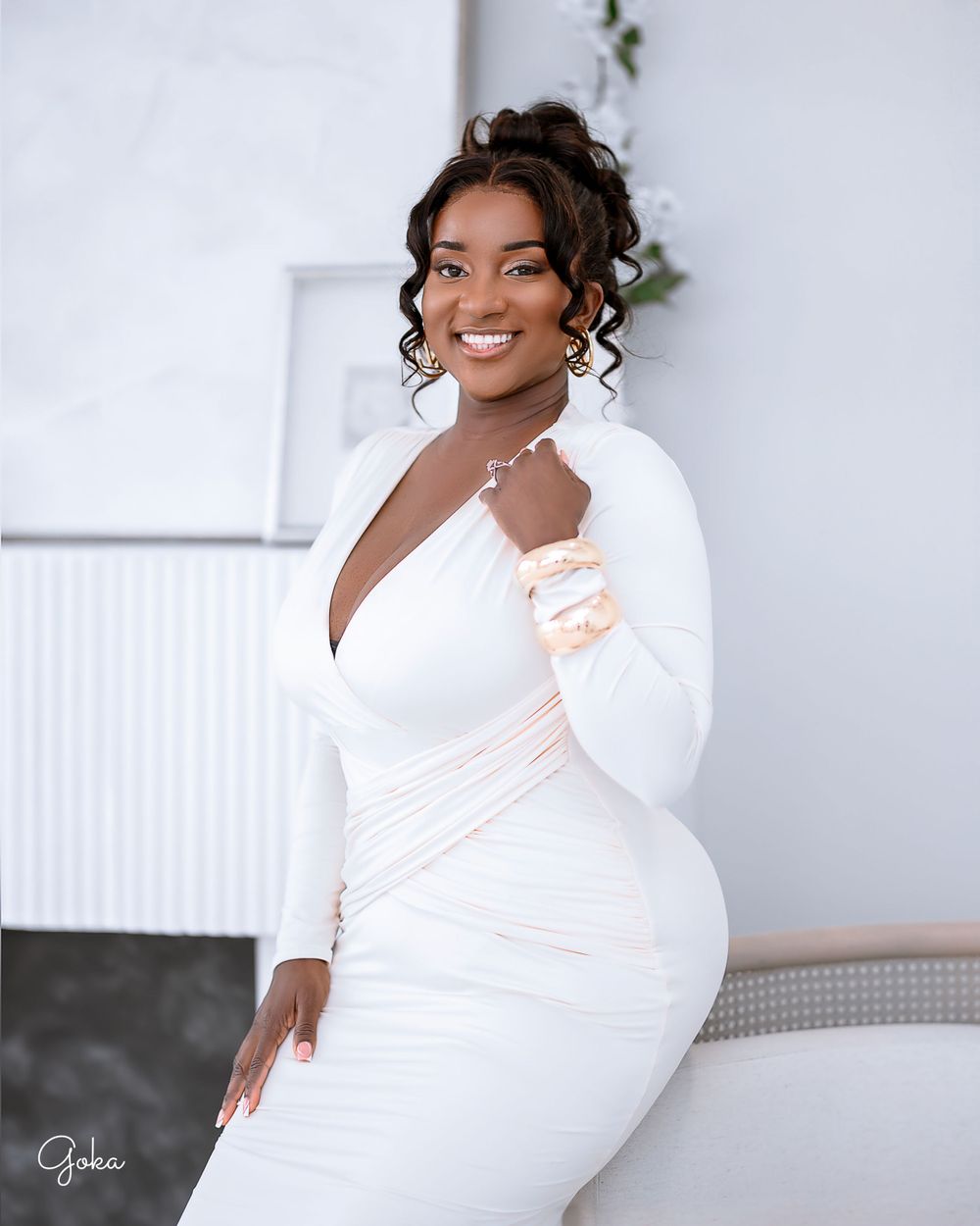 Someone wearing an elegant form-fitting white dress with long sleeves poses against a light-colored wall with decorative greenery.