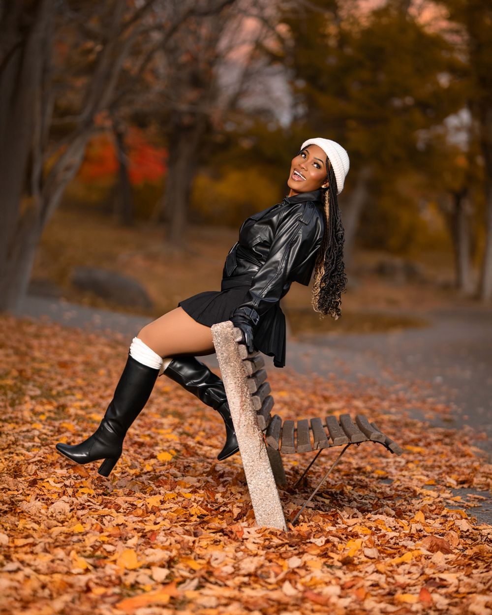 Fashionable model in leather outfit and white beanie poses on wooden bench surrounded by autumn leaves in park.