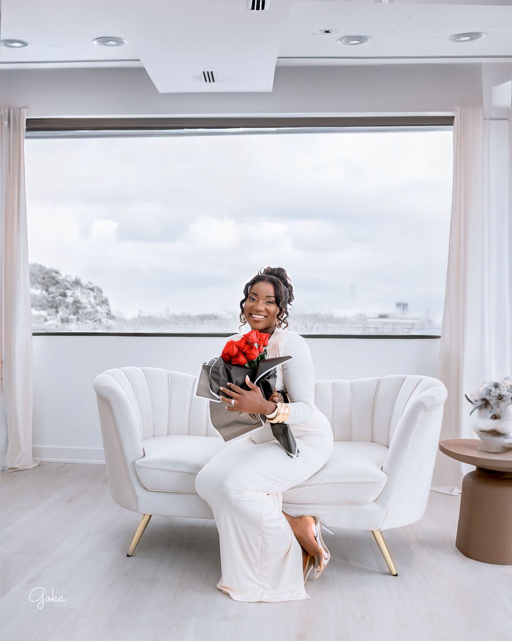 A person in white attire sits on a modern white armchair holding a small black dog with a red bow.