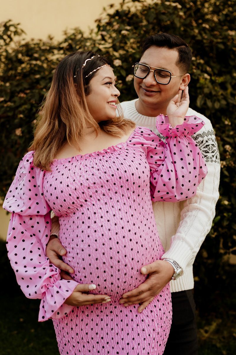 Expecting couple poses outdoors, with woman wearing pink polka dot maternity dress and man in white sweater.