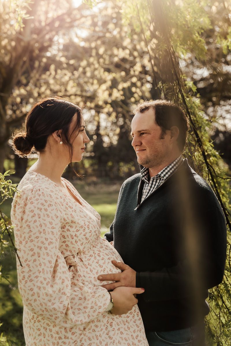 Intimate maternity photo of couple in garden with sunlight filtering through trees.