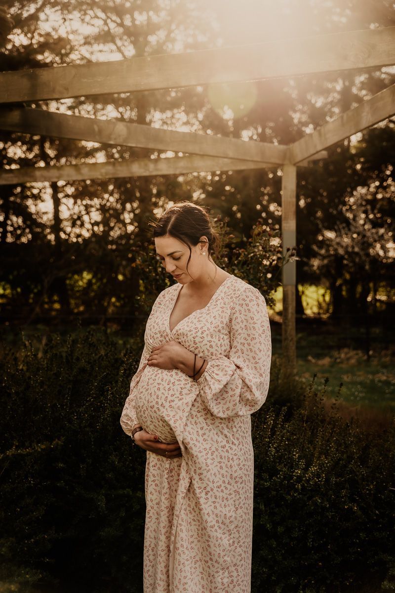 Serene maternity portrait of woman in floral dress under wooden pergola at golden hour.
