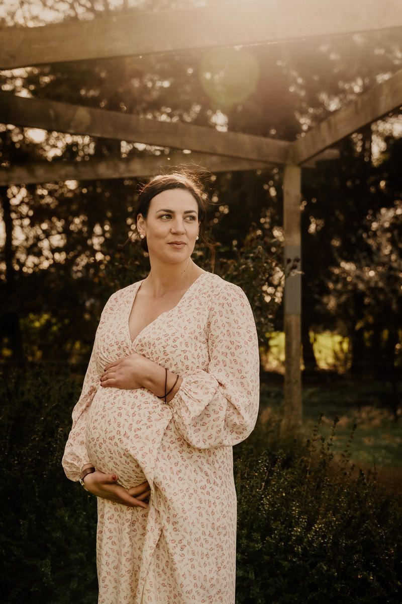 Maternity portrait of woman in floral dress standing under wooden pergola at sunset.
