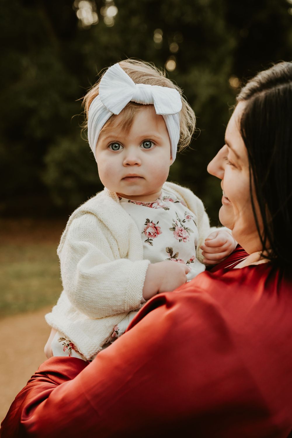 Baby wearing white bow headband and floral outfit being held outdoors.