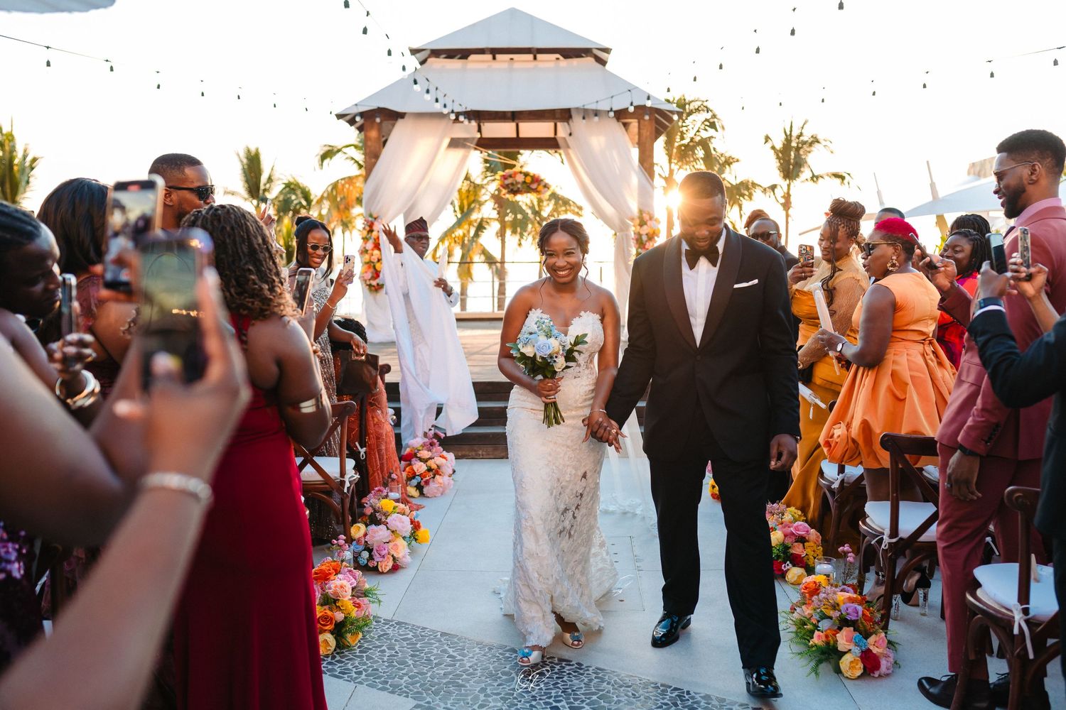 A joyous wedding ceremony takes place at a beachfront gazebo decorated with vibrant tropical flowers at sunset.