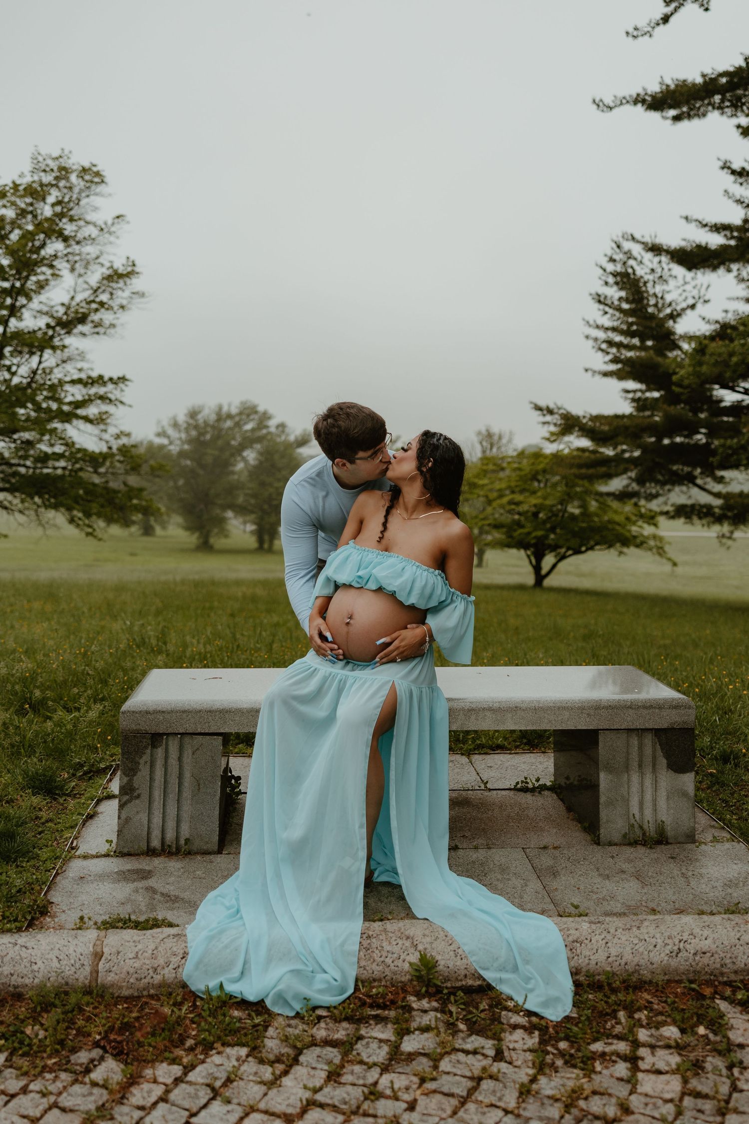 Romantic maternity photoshoot in a park with couple in matching light blue outfits sitting on stone bench.