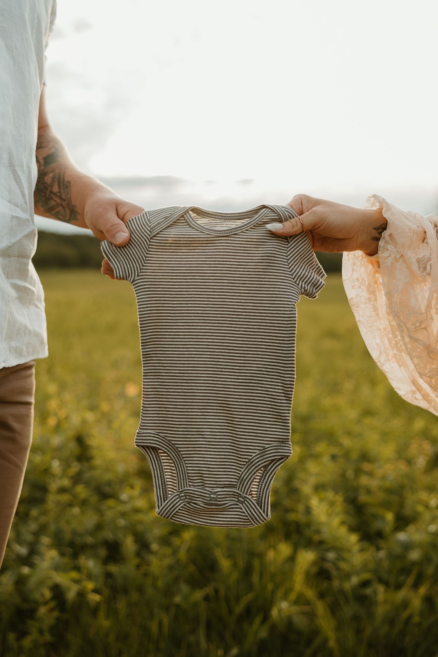 A striped baby onesie held up against a sunny outdoor background with green grass and natural lighting.