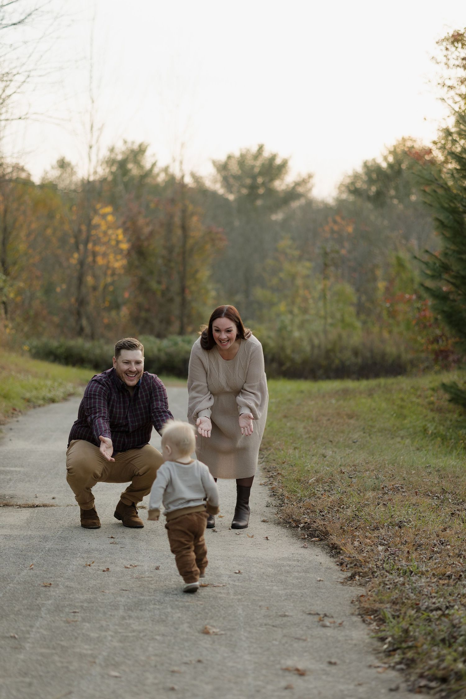 A toddler takes first steps on a path while supported by adults during an autumn outdoor photo session.