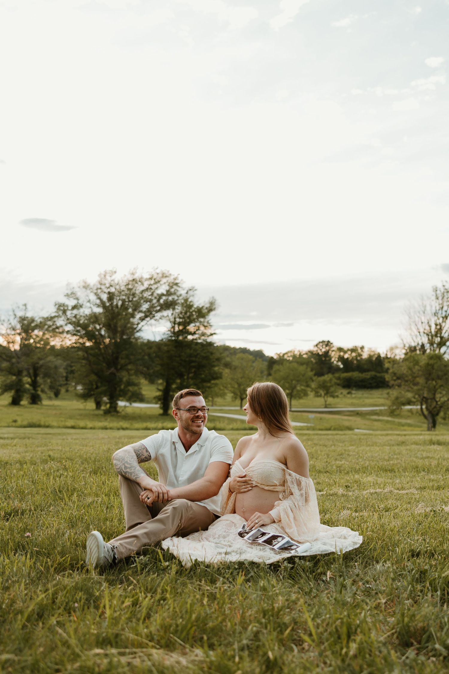 A couple sits together on a blanket in a grassy meadow at sunset enjoying a peaceful moment outdoors.