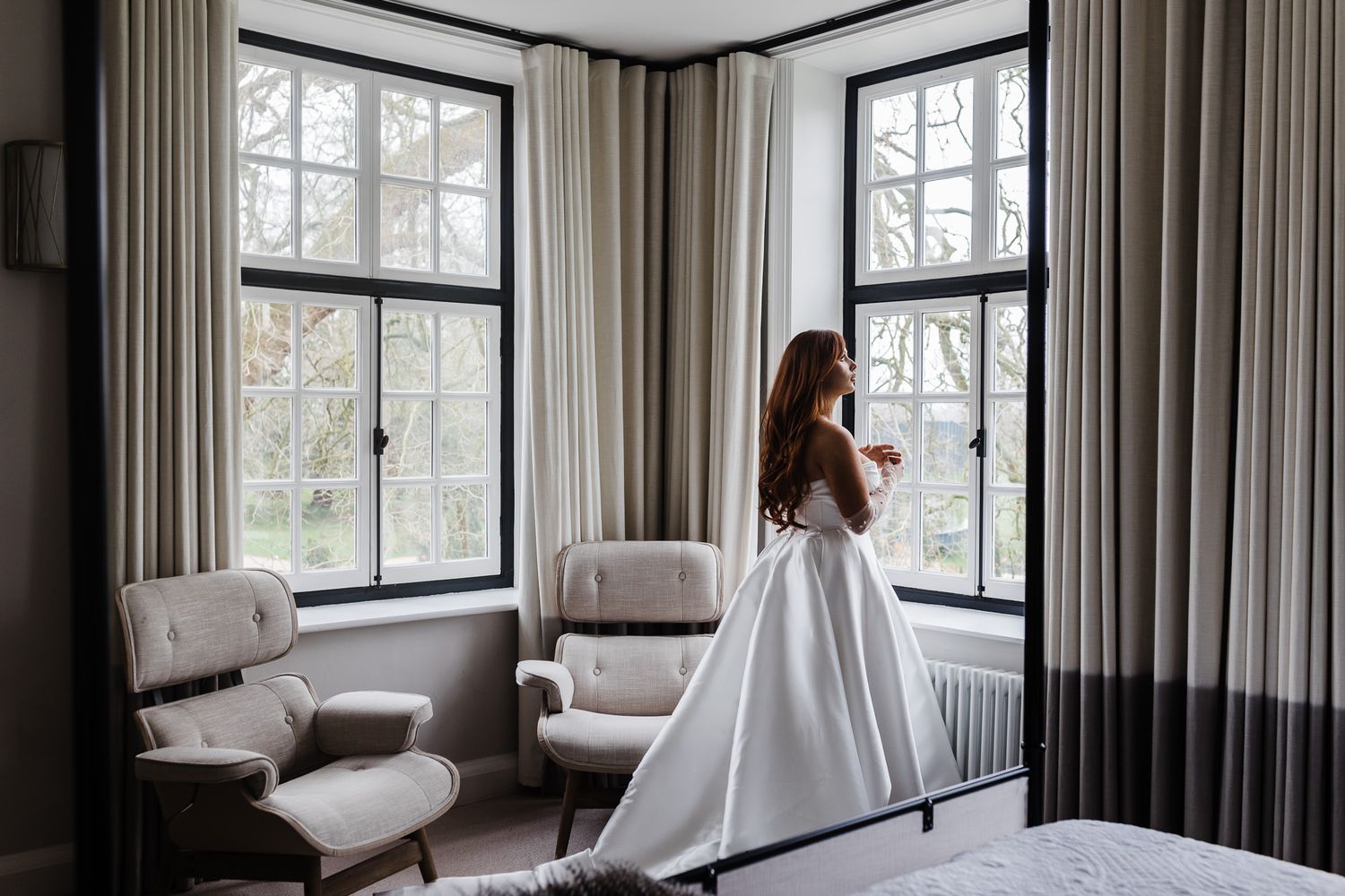 A bridal silhouette against tall windows in an elegant room with beige curtains and white armchairs. Sant Ffraed House Wedding Photographer