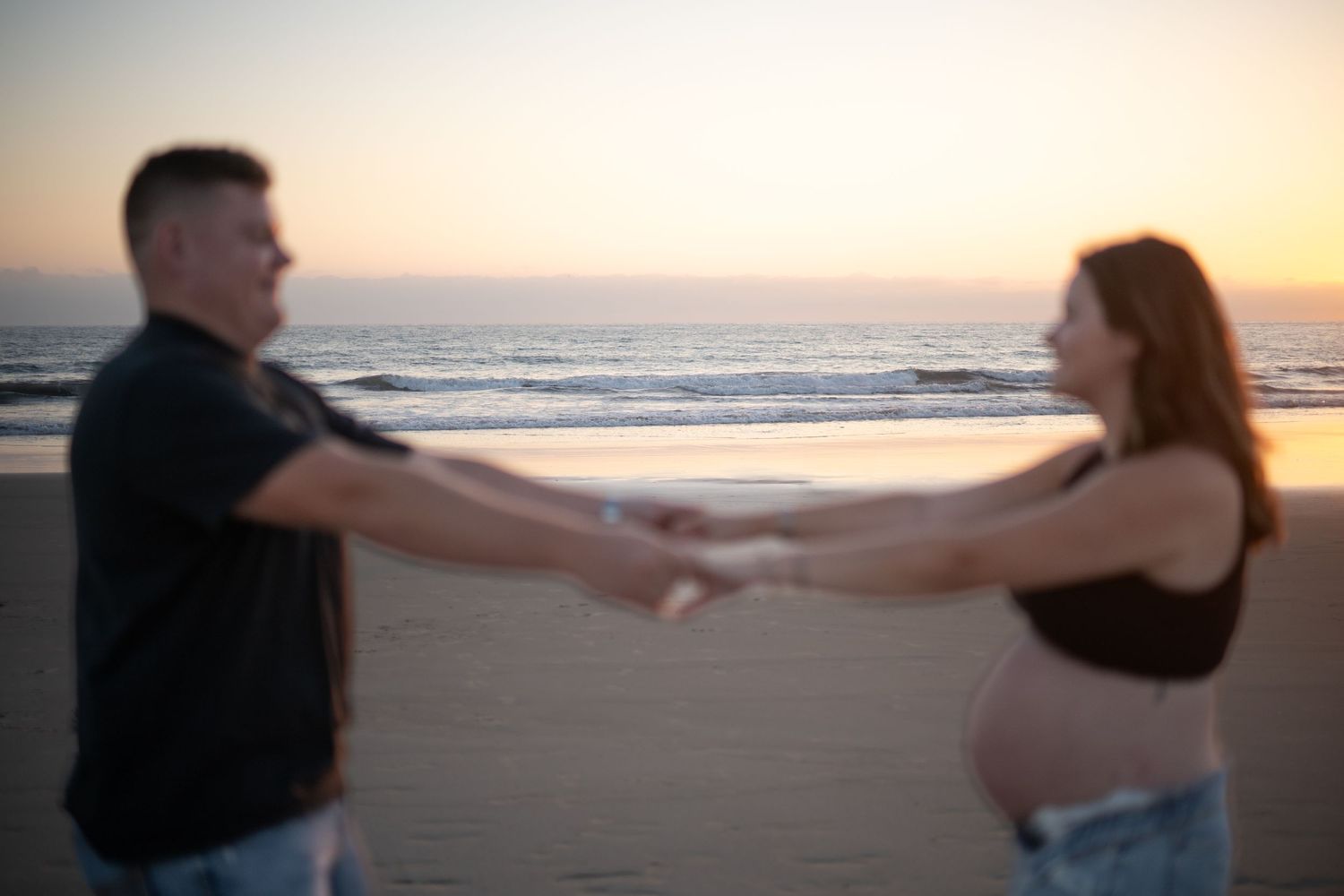 Couple holding hands while walking on beach at sunset, ocean waves crashing in background.