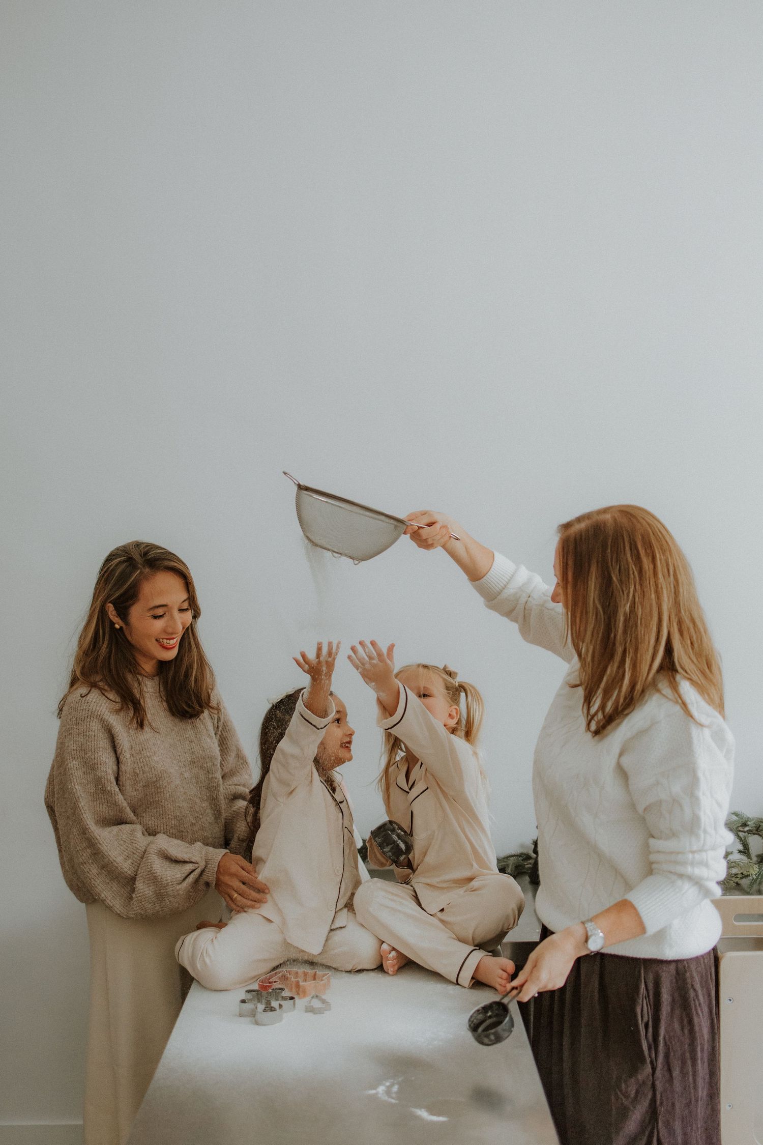 Two women and two children playing joyfully with confetti and a strainer in a bright white room.