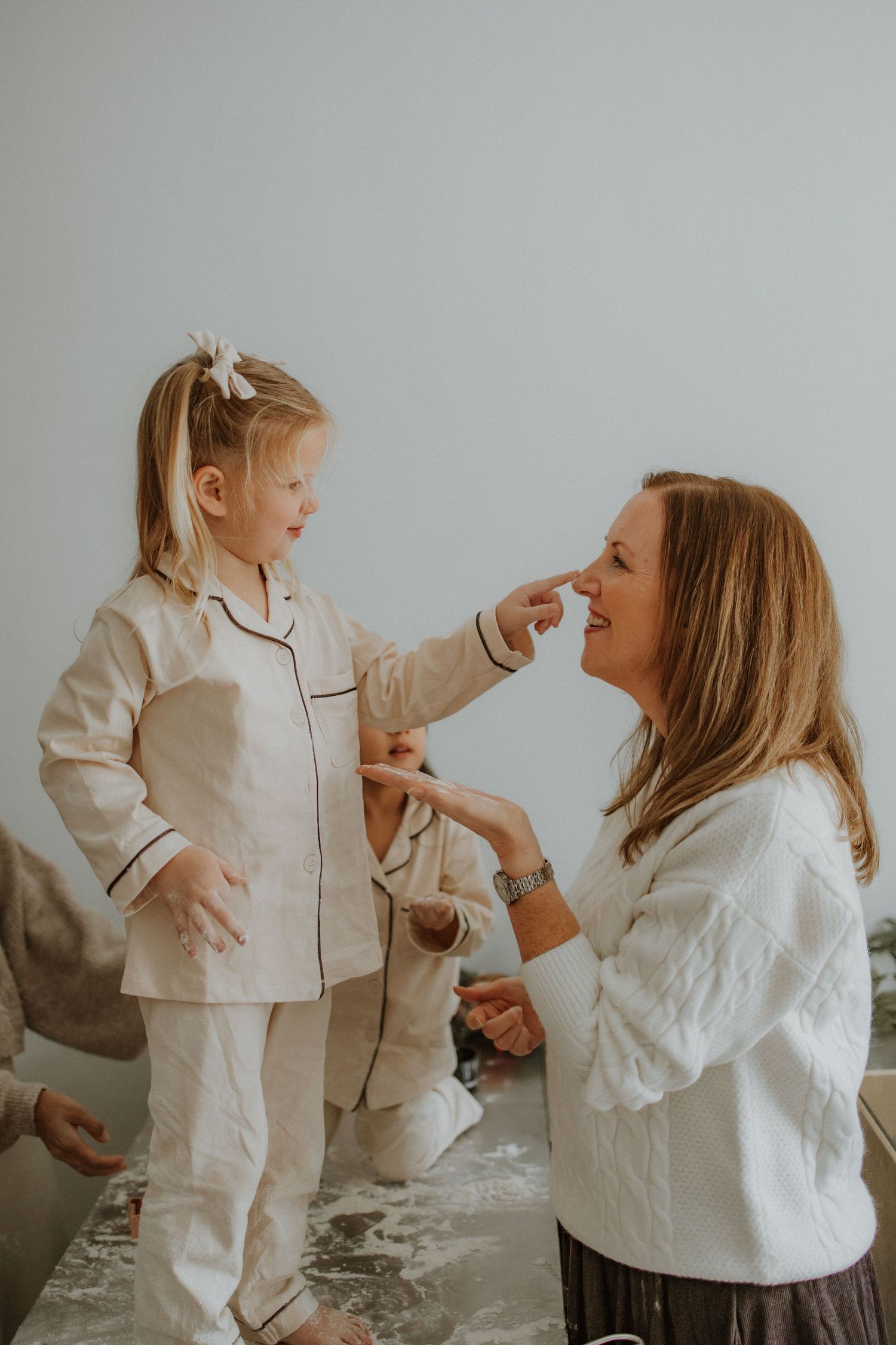 A young girl in cream pajamas playfully touches a smiling woman's nose while baking together in a bright kitchen.