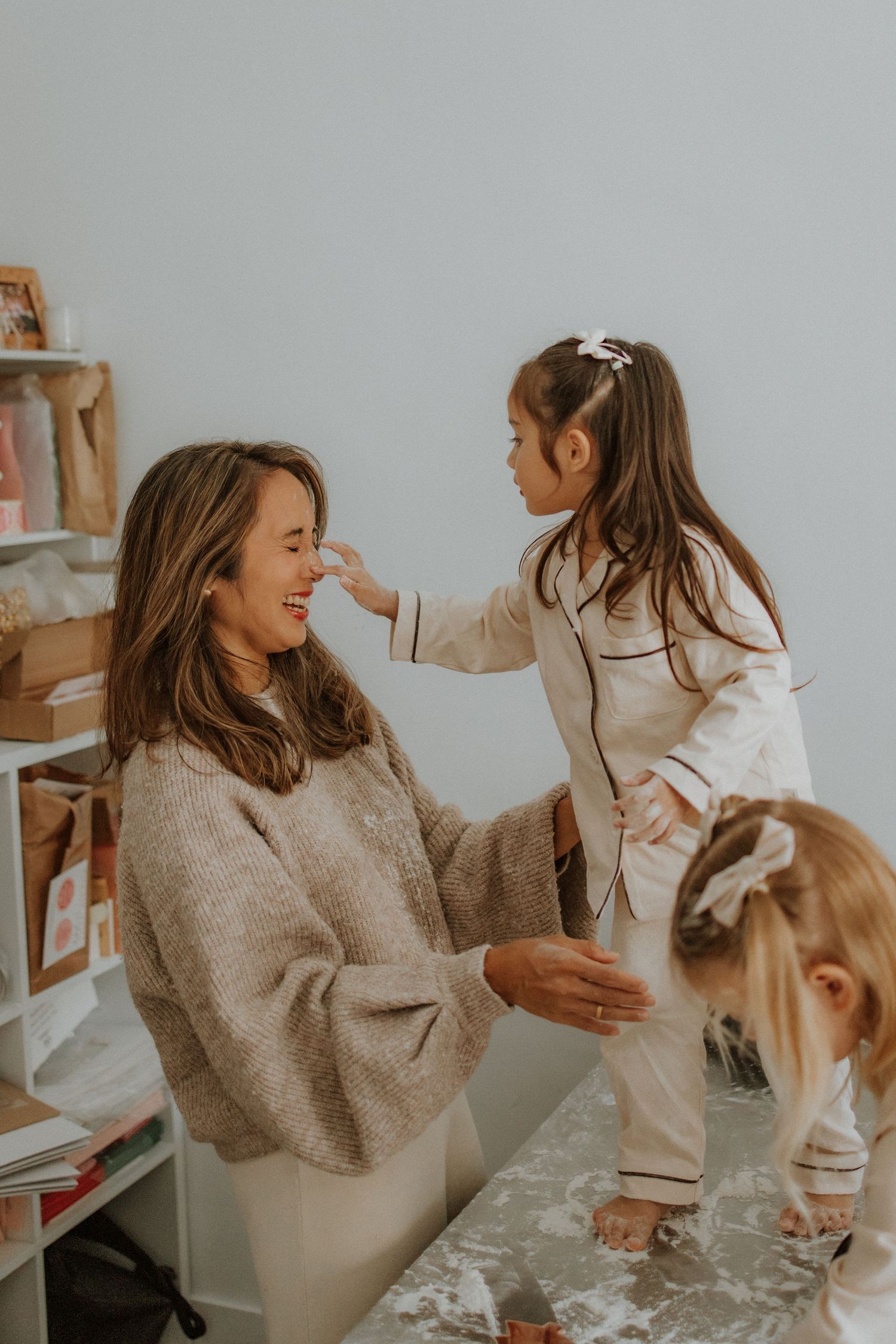 A smiling mother in a beige sweater plays with two young girls in white pajamas in a bright, cozy room.