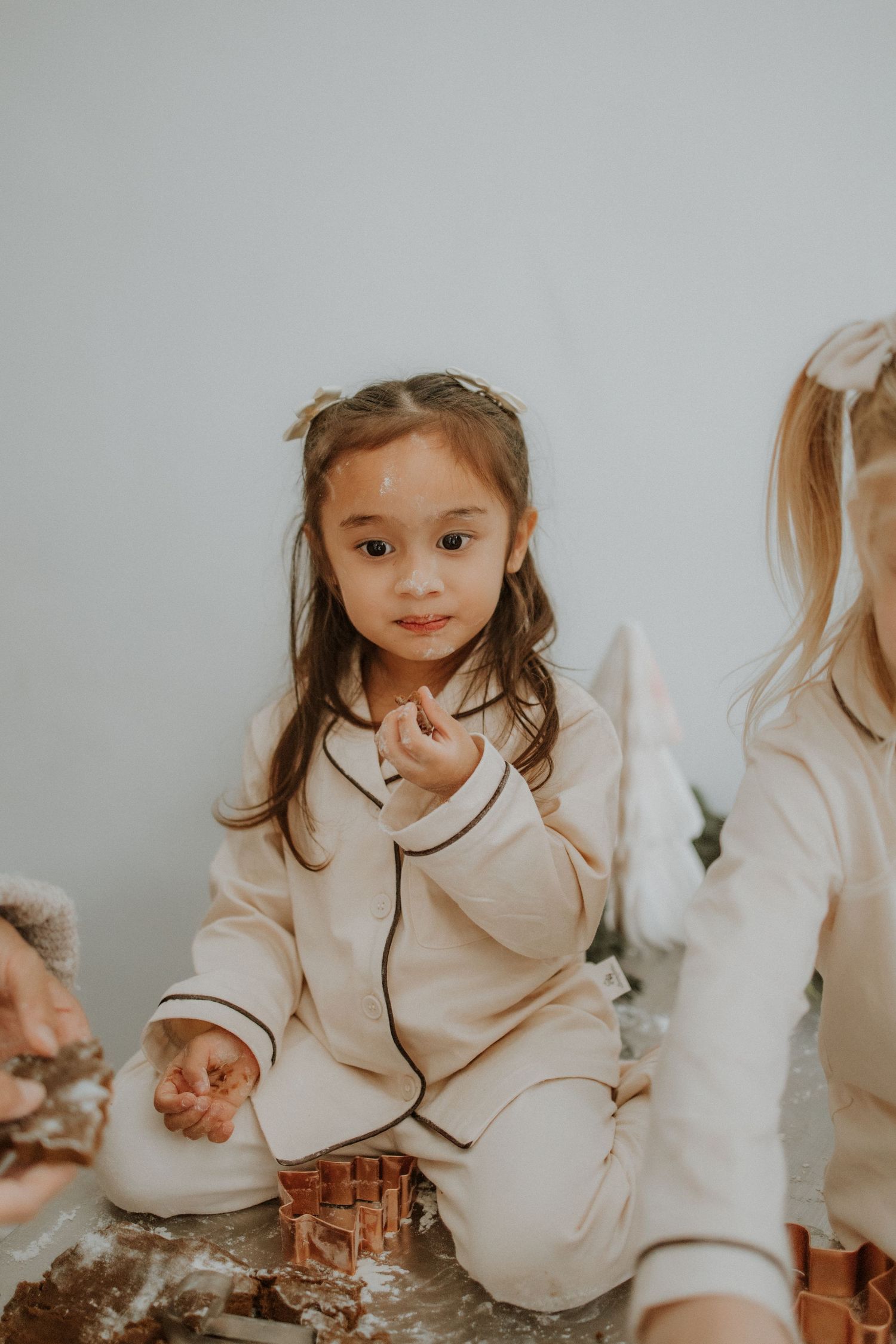 Young girl in cream pajamas sits on floor dusted with flour, looking at camera during holiday baking.