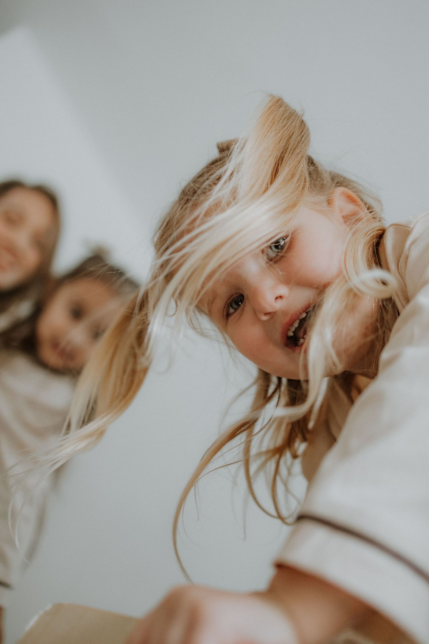 Playful blonde girl tilting her head toward camera with hair flying, two children visible in soft white background.