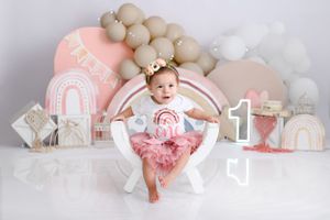 First birthday photo setup with pink decor, balloons, and an elegant white chair against a neutral backdrop.