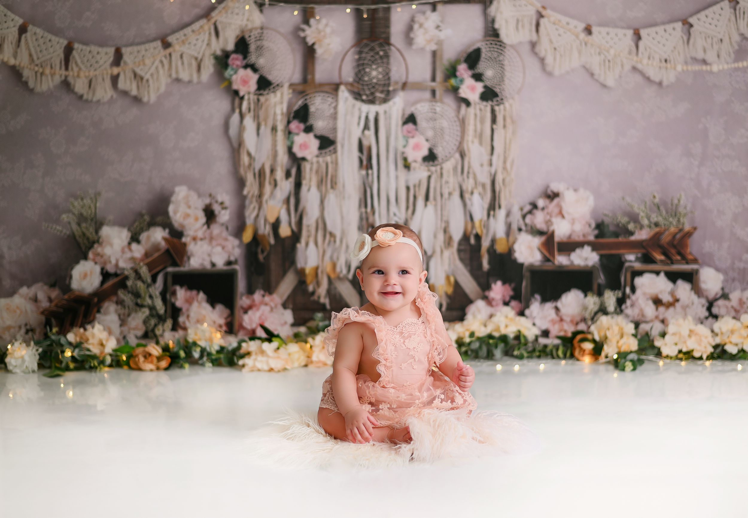 A baby in a pink dress sits on a white floor with floral decorations and banners hanging in the backdrop.