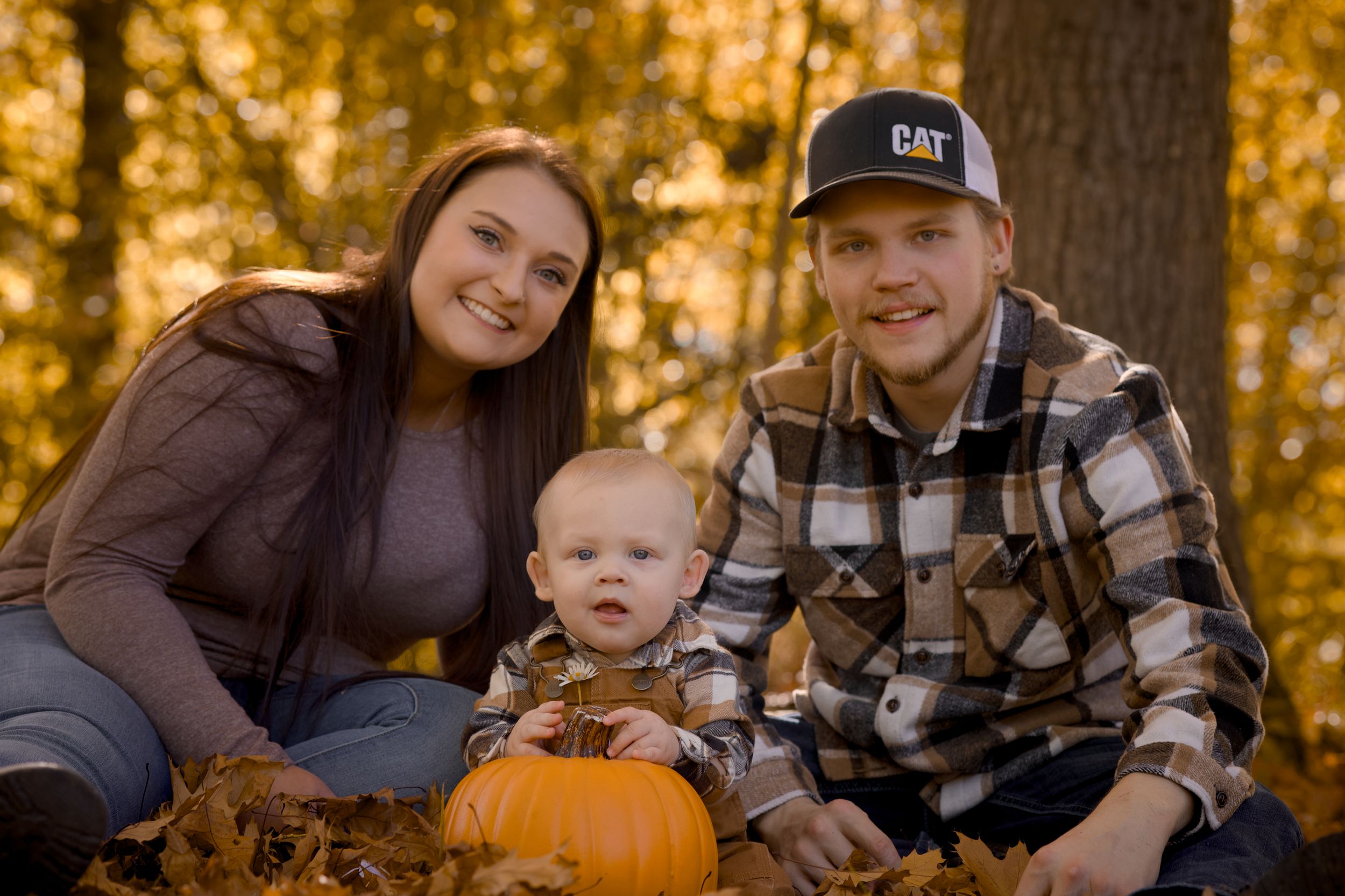 A family sits together on fallen autumn leaves with a pumpkin during a golden sunset photo session in the woods.