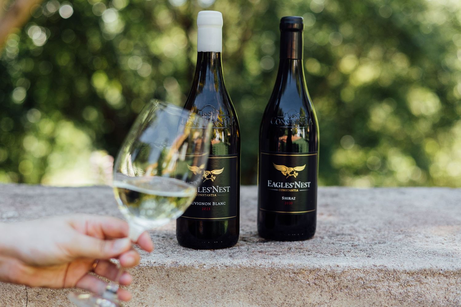 Two bottles of Eagle's Nest wine with a hand holding a glass of white wine against a blurred green outdoor background.