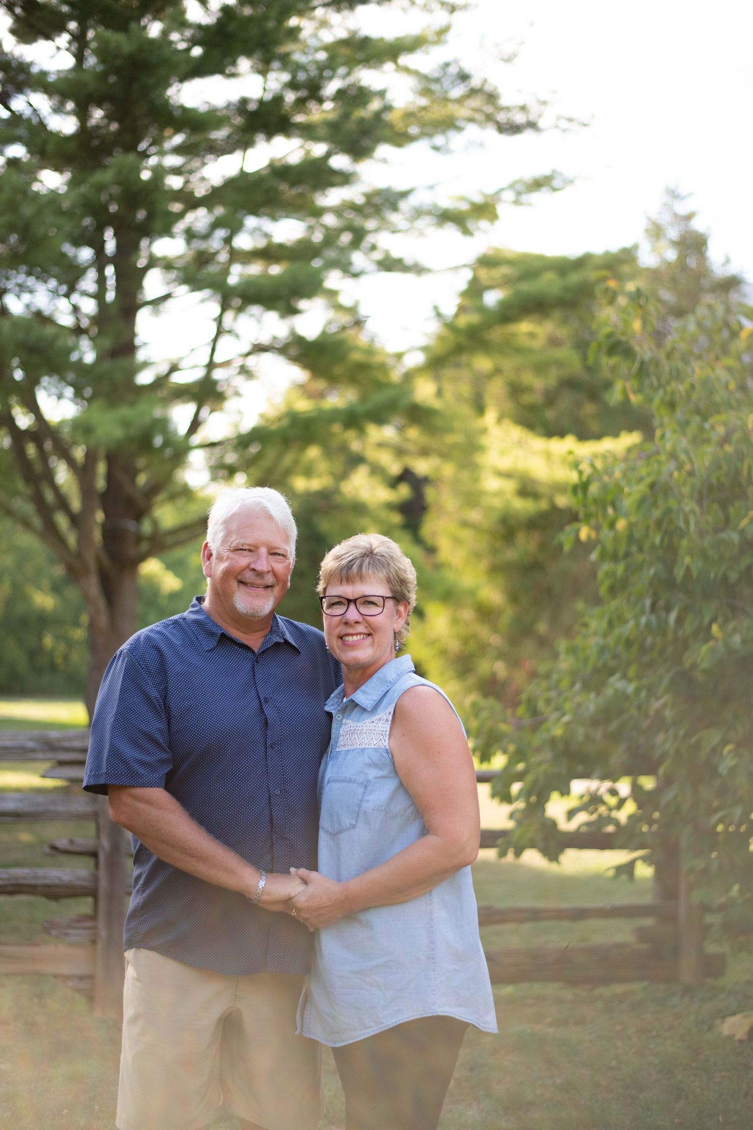 A couple embraces while posing outdoors near a wooden fence with green trees in the background during golden hour.