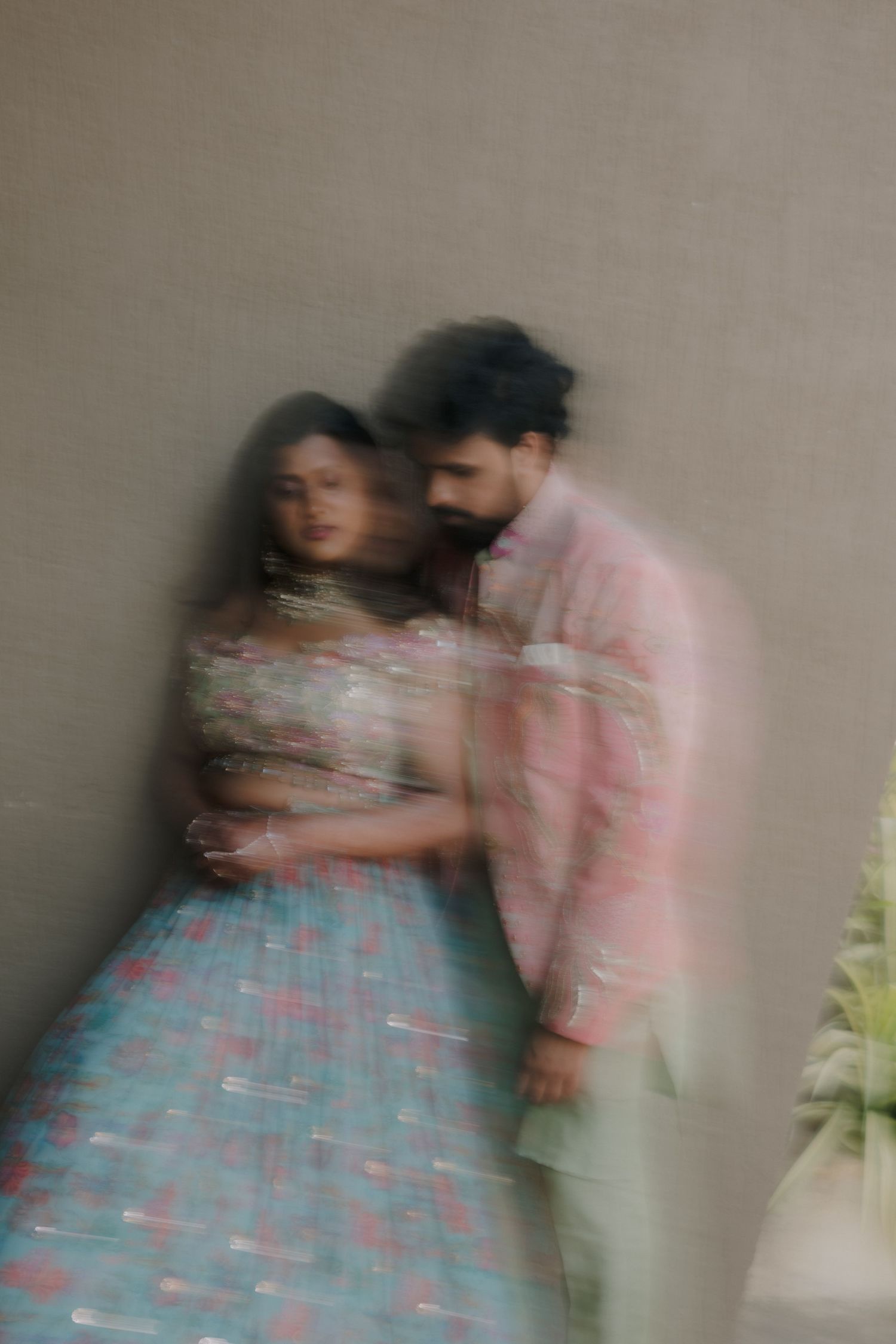Couple in traditional Indian attire embrace tenderly against a neutral wall in a dreamy motion-blur photo.