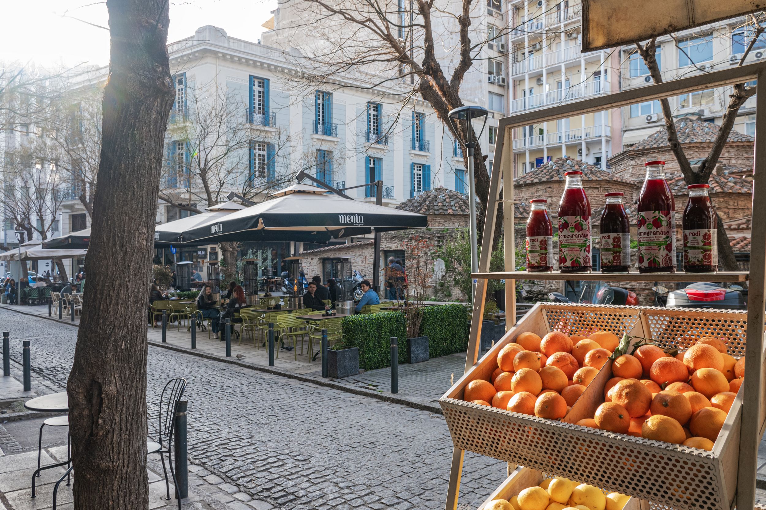 Outdoor market stall with fresh oranges and colorful juice bottles, with a bustling café terrace in the background.