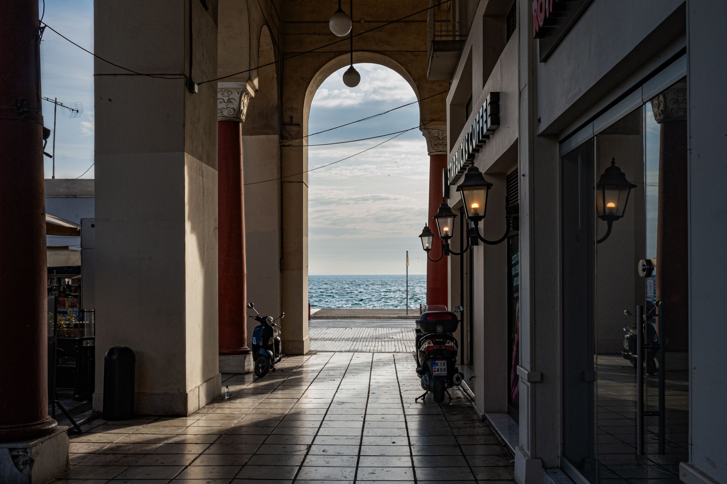 A scenic arcade walkway with arched columns frames a stunning ocean view at dusk, with scooters parked along the corridor.