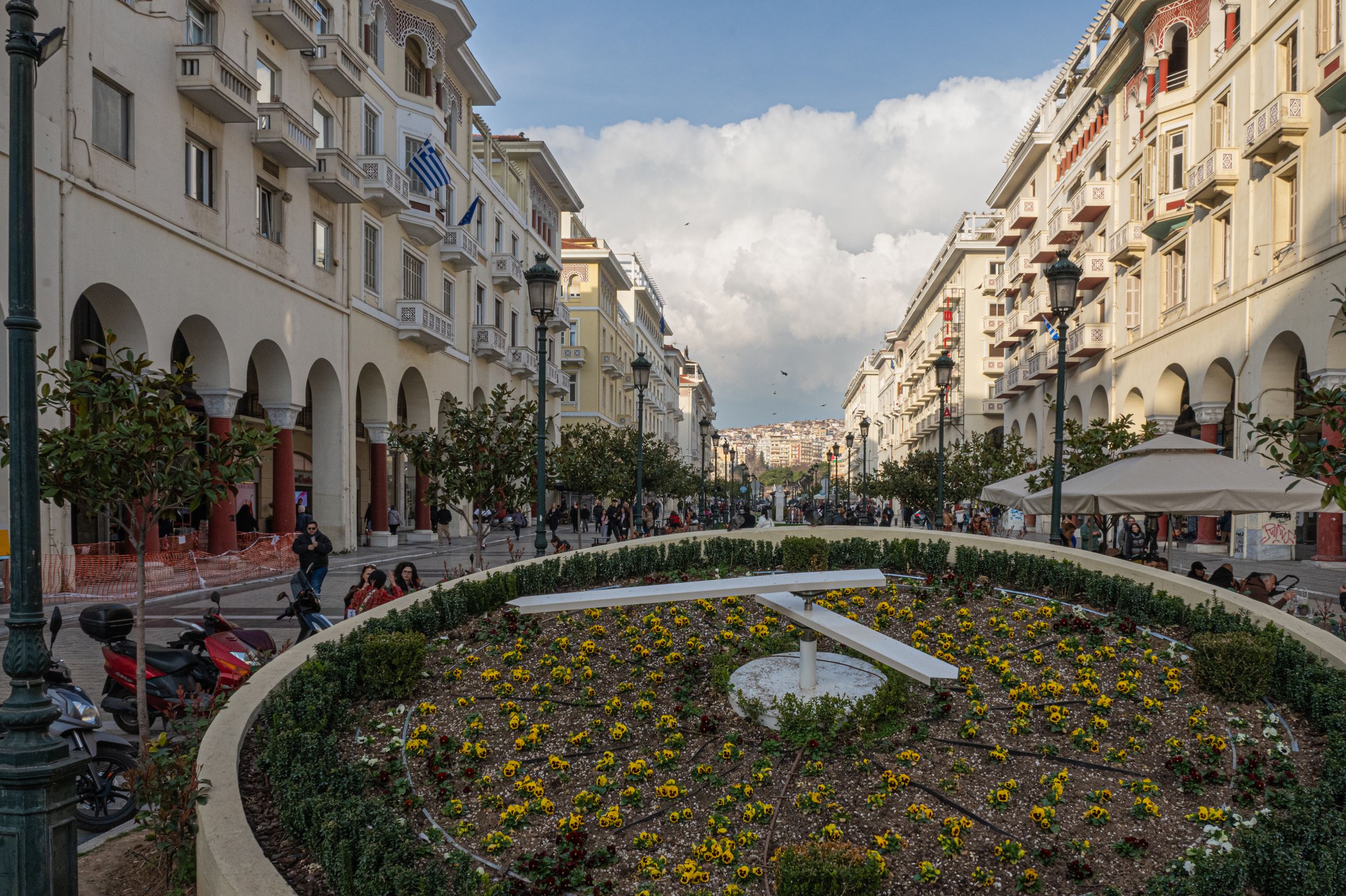 Aristotelous Square in Thessaloniki, Greece, featuring colorful flower gardens and neoclassical buildings lining a pedestrian boulevard.