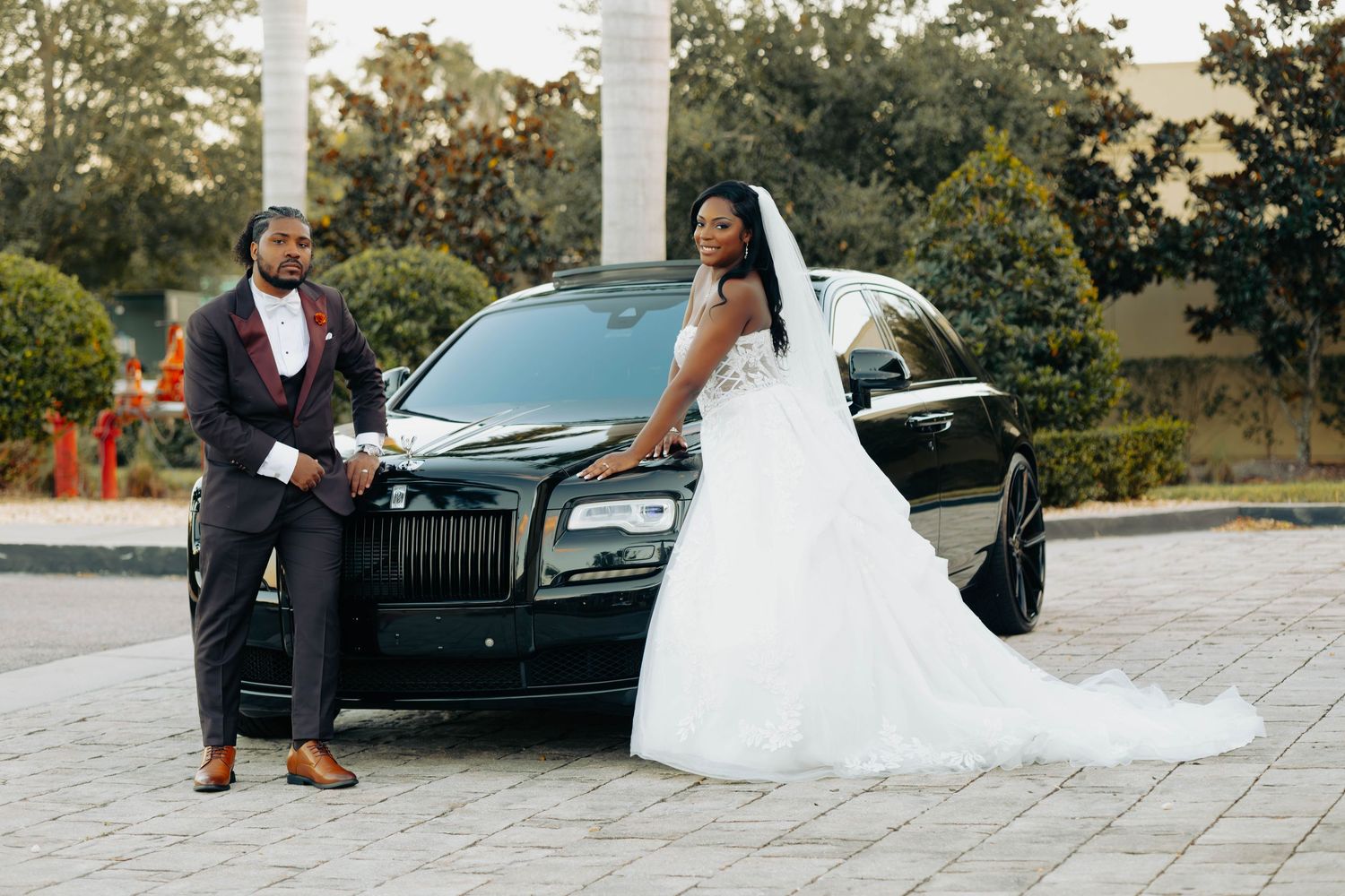 Bride and groom posing elegantly in front of a black Rolls-Royce on their wedding day.