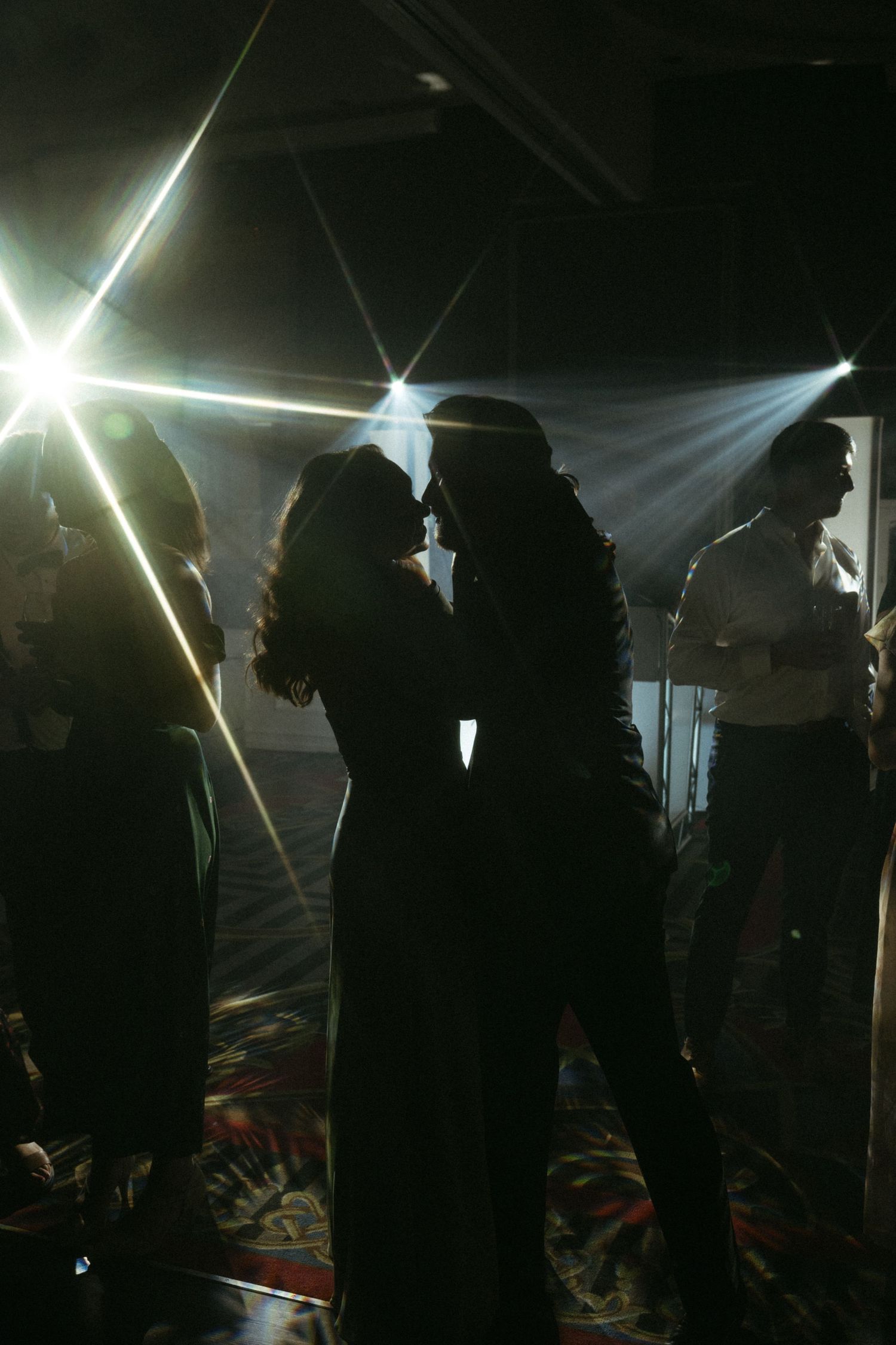 Two silhouetted figures slow dancing at a dimly lit event with bright stage lights beaming overhead.