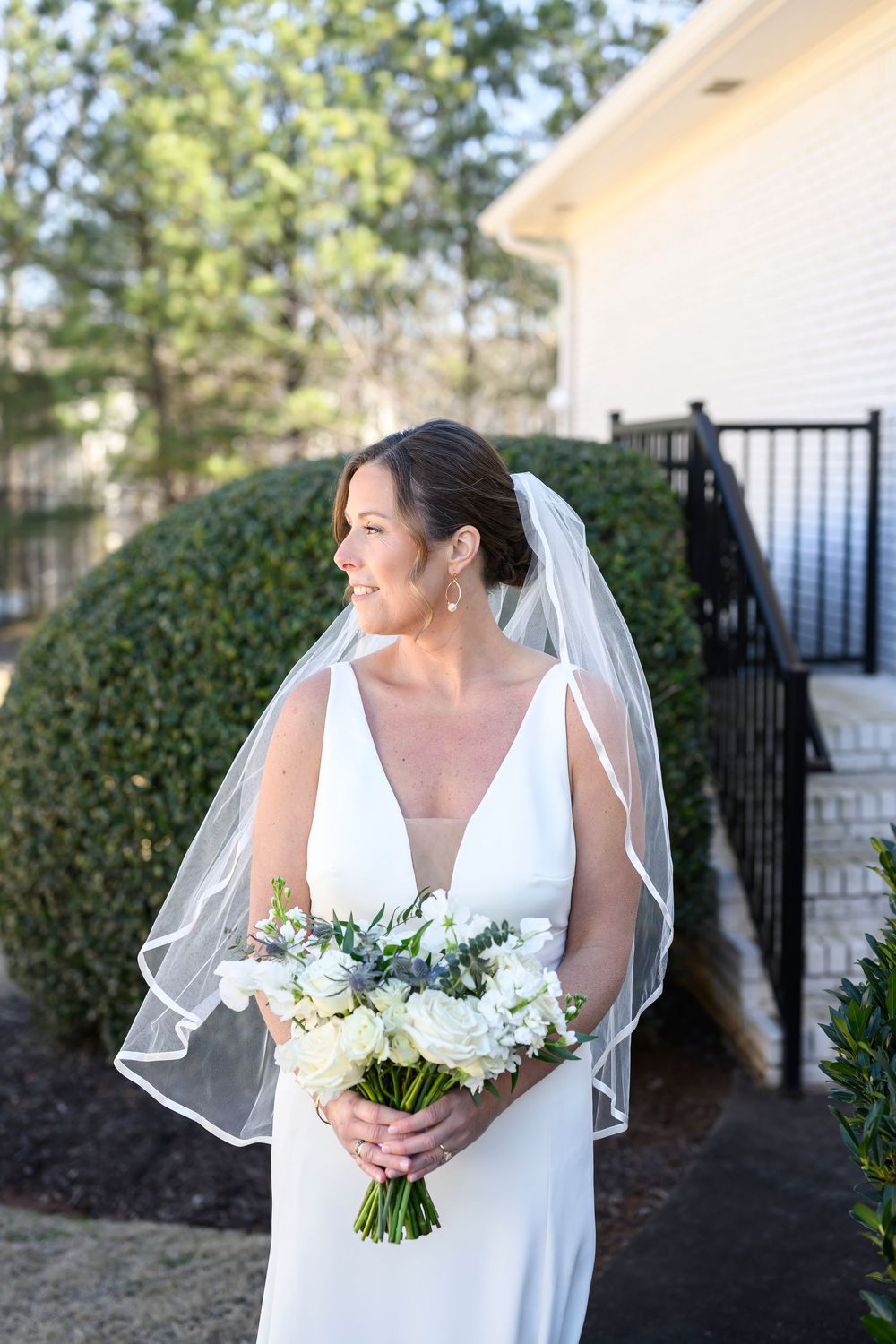 A bride in a classic white wedding dress holds a delicate bouquet of white and green flowers against an outdoor backdrop.