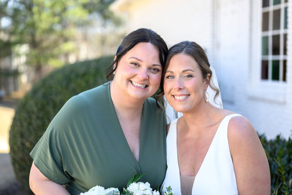 Two people wearing green and white outfits smile together while holding white floral bouquets outdoors.