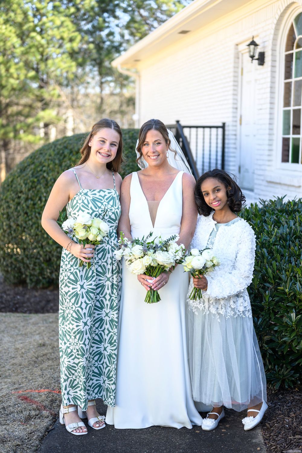 Three people in formal attire pose together outside a white church building holding white floral bouquets.
