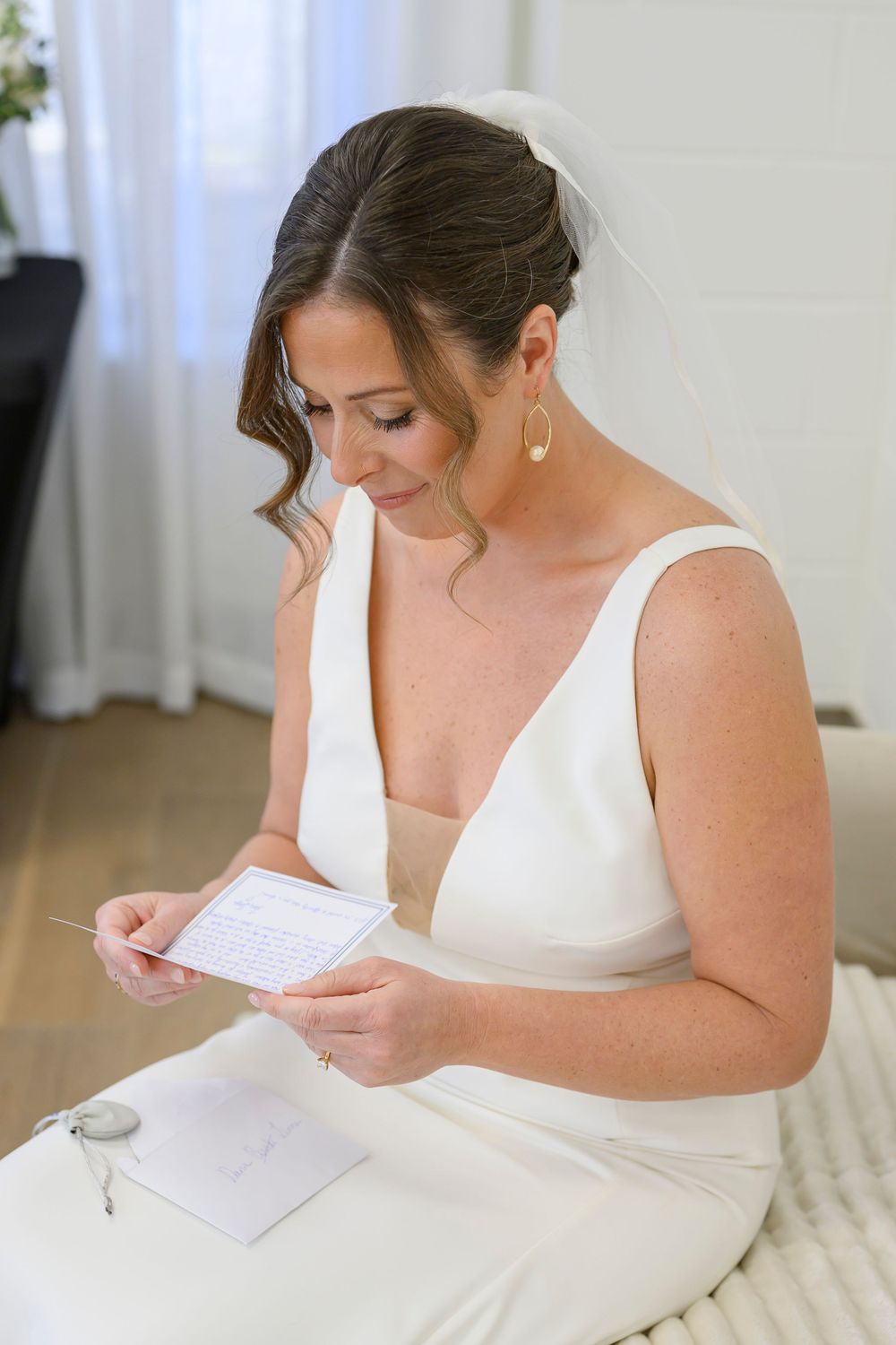 A bride in an elegant white dress sits reading a letter on her wedding day, wearing pearl earrings and an upswept hairstyle.