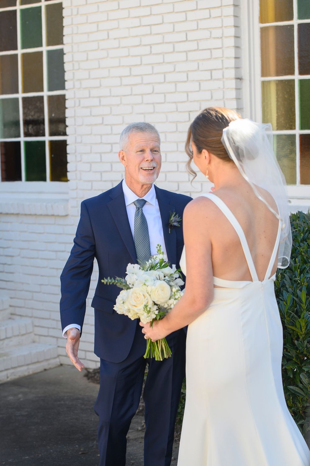 A touching first look moment between a bride in a white dress and someone in a navy suit outside a white brick building.